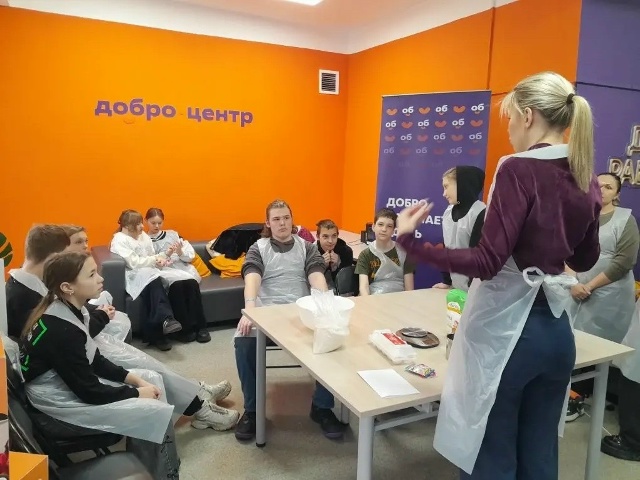 На фотографии изображено занятие. Группа детей сидит полукругом возле стола, на котором лежат различные материалы. Все дети одеты в фартуки, что предполагает участие в практической деятельности, связанной с кулинарией. Ведущий занятия - женщина стоит справа. Она активно жестикулирует. Стены помещения окрашены в яркие цвета: одна стена оранжевая с надписью "добро центр", а другая - фиолетовая.